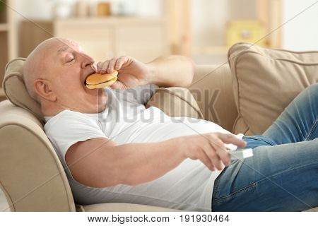 Fat senior man watching TV and eating hamburger while lying on sofa at home. Sedentary lifestyle concept