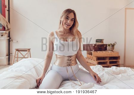 Sexy blonde woman with big breasts in a white top and corset sits on the bed.