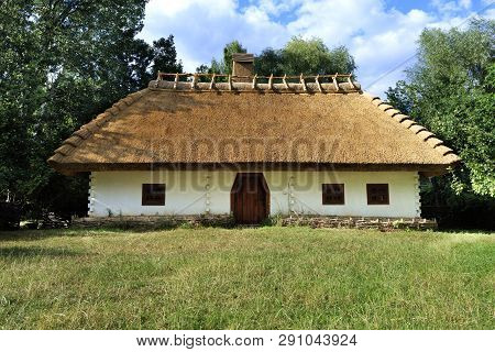 Old Traditional Ukrainian Rural House With A Thatched Roof And A Wicker Fence In The Garden With Gre