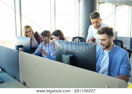 Group Of Young People In Casual Wear Sitting At The Office Desk And Discussing Something While Looki