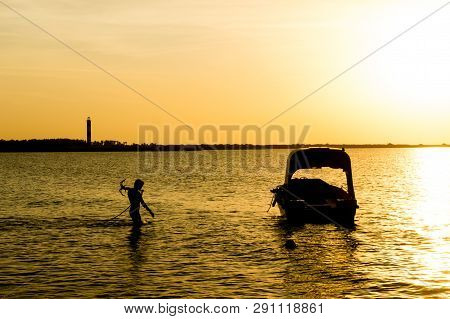 Silhouette Of A Man Taking Anchor For A Boat With Setting Sun In The Distance