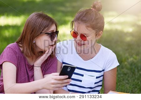 Image Of Two Girls Looking At Smart Phone And Talking In Park. Attractive Females Look Thoughtful Si