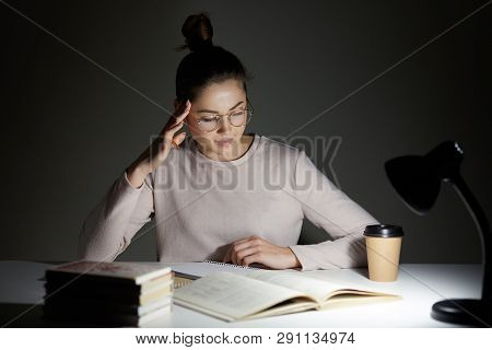 Beautiful Student Sits At White Desk Against Dark Background, Tries To Concentrate Under Light Of La