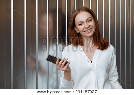 Satisfied Female Entrepeneur With Toothy Smile, Wears White Jumper, Holds Modern Cell Phone, Sends T