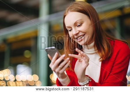 Positive Pleased Female Model Uses Mobile Phone Data, Uses Chat Application, Holds Modern Cell Phone