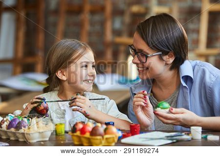 Portrait Of Happy Little Girl Painting Easter Eggs With Mothera Nd Looking At Each Other