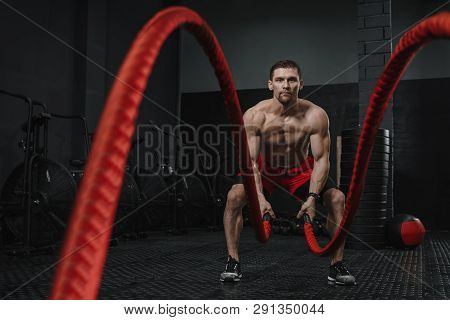 Muscular Man Doing Battle Ropes Exercise At The Gym. Athlete Training With Red Rope. The Sport Motiv
