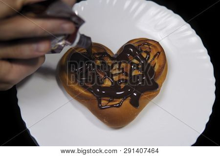 Someone Pours Chocolate On A Sweet Heart-shaped Bun, Fresh Baking Lies On A White Plate On A Black B