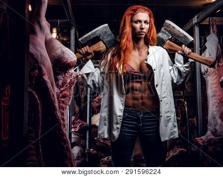 Strong Fitness Woman Wearing Unbuttoned White Shirt Posing With Two Axes In A Refrigerated Warehouse