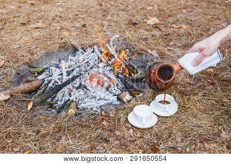 A Man Pours Coffee From An Earthen Turkish Pot Into Small White Cups On A Campfire In The Spring For