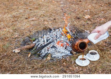 A Man Pours Coffee From An Earthen Turkish Pot Into Small White Cups On A Campfire In The Spring For
