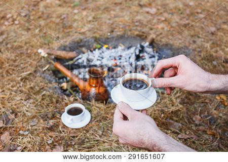 A Man Pours Coffee From An Earthen Turkish Pot Into Small White Cups On A Campfire In The Spring For