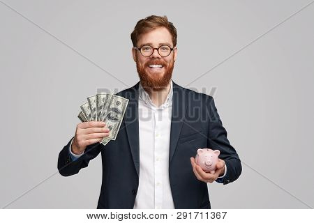 Handsome Businessman With Ginger Beard Smiling And Holding Piggy Bank And Bunch Of Dollar Bills Whil