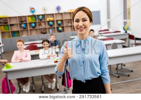 Laughing Teacher In Blue Blouse Standing In Front Of Pupils And Showing Thumb Up