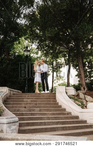 Beautiful Cuple Stand On The Stairs And Hug Each Other. Man And Woman Walking In The Park. Love Stor