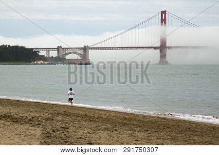 Person Jogging On Beach In Front Of Golden Gate Bridge, San Francisco, California, Usa. Bridge Is Pa
