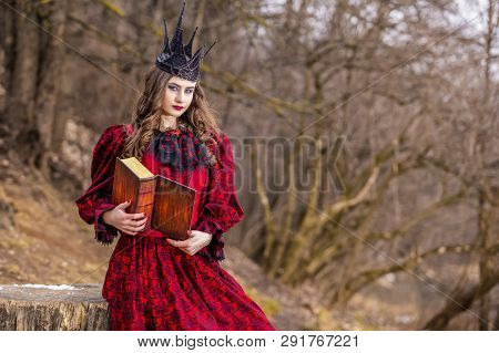 Art Photography. Mysterious Medieval Queen In Red Dress And Spiky Black Crown Posing With Ancient Bo