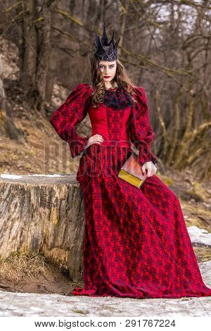 Art Photography. Mysterious Medieval Queen In Red Dress And Spiky Black Crown Posing With Ancient Bo