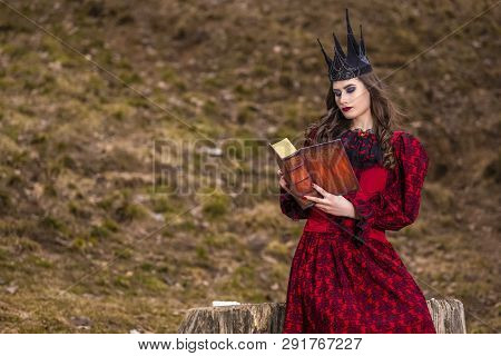 Art Photography. Mysterious Fairy Medieval Queen In Red Dress And Spiky Black Crown Posing With Anci