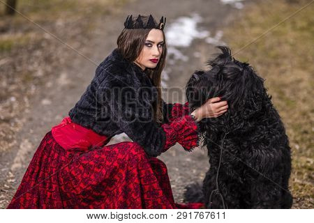 Beautiful Princess In Red Dress And Black Fur With Crown And Her Dog In Forest During Early Spring. 