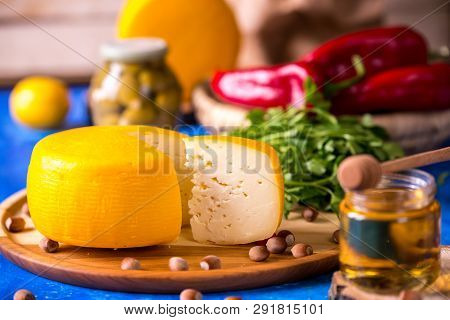 Cheese Wheel On A Wooden Board Lies On The Table With Food And Fresh Herbs. Blurred Background.