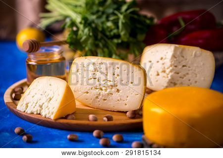 Cheese Wheel On A Wooden Board Lies On The Table With Food And Fresh Herbs. Blurred Background.