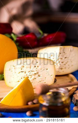 Cheese Wheel On A Wooden Board Lies On The Table With Food And Fresh Herbs. Blurred Background.