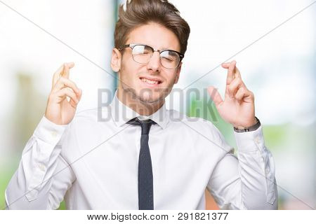 Young business man wearing glasses over isolated background smiling crossing fingers with hope and eyes closed. Luck and superstitious concept.