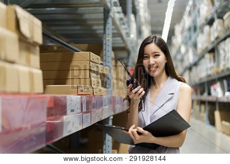 Smart Smiling Young Asian Woman Working In Store Warehouse. She Is Standing And Holding Document Fol