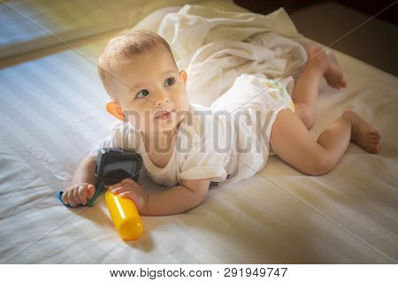 Little Baby 8 Months Lies On A White Bed. Infant Girl Plays With The Action Camera And Looking At Mo
