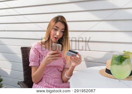 Surprised Woman By Invoice, By Phone. The Girl In Summer Cafe On Veranda Of Restaurant. Sits At A Ta