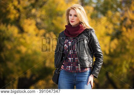 Young fashion blonde woman walking in city park  Stylish female model in black leather jacket and snood scarf