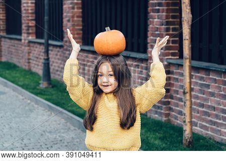 Funny Child Girl In Orange Pullover For Halloween With Pumpkin And On A Dark Brick Background. Happy