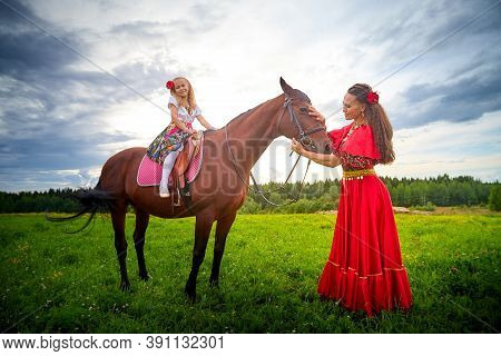 A Woman And A Girl In Bright Gypsy Dresses With A Horse In A Field With Green Grass. Mother And Daug