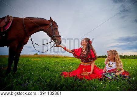A Woman And A Girl In Bright Gypsy Dresses With A Horse In A Field With Green Grass. Mother And Daug