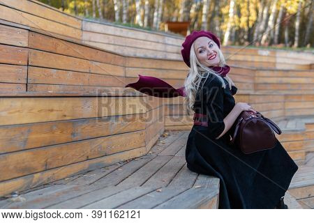 Smiling Woman Sits On A Bench Of A Summer Theater, Wooden In A Burgundy Coat And Biret, In The Fall 