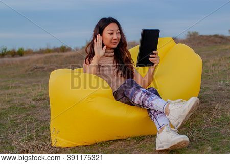 Pretty Young Asian Woman Sitting On A Yellow Lamzak Having A Video Chatting With Tablet On A Cliff N