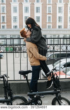 Loving Young Man Holding His Girl In The Air By Her Thighs. Side View