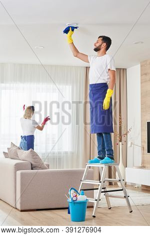 Man Cleaning Ceiling And Lamps In Living Room. House Cleaning Service Concept.