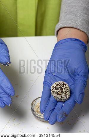 A Woman In Rubber Gloves Makes Candies From Dried Fruits And Nuts. Some Are Sprinkled With Sesame Se