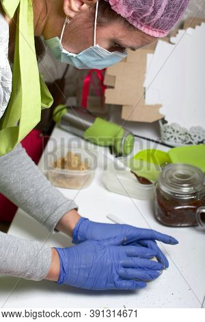 A Woman In Rubber Gloves And A Mask Makes Candies From Dried Fruits And Nuts At Home In Self-isolati
