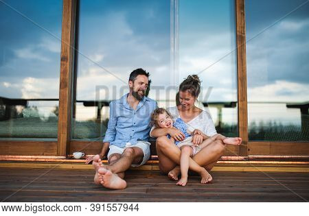 Family With Small Daughter Sitting On Patio Of Wooden Cabin, Holiday In Nature Concept.