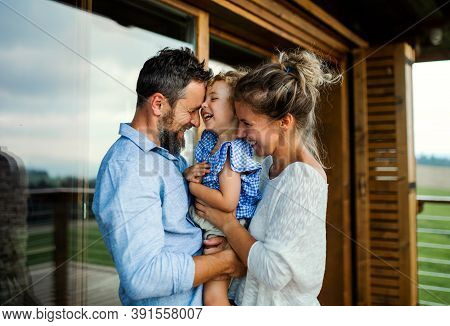 Family With Small Daughter Standing On Patio Of Wooden Cabin, Holiday In Nature Concept.