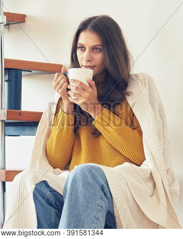 Bautiful Young Woman Sitting Onstairs In Warm Clothes And Drinking Coffee, Wrapped In Blanket
