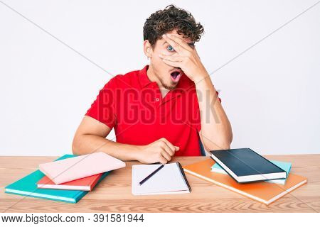 Young caucasian man with curly hair sitting on the table stuying for university peeking in shock covering face and eyes with hand, looking through fingers afraid 