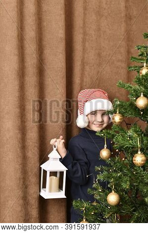 A Child With A Lantern In His Hand Stands Embarrassedly Smiling Near A Dressed Christmas Tree.