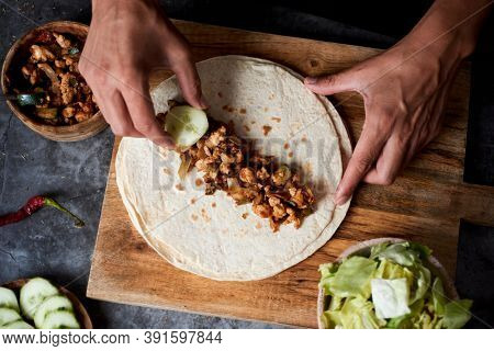 high angle view of a young caucasian man preparing a durum or a burrito, with chicken meat cooked with different vegetables such as onion or red and green pepper, and fresh lettuce and raw cucumber