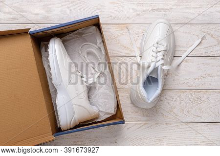 One White Chunky Sole Sneakers In A Brown Cardboard Box, The Other On The White Wood Floor. Open Box