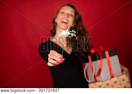 Happy Holiday Celebration Concept. Laughing Woman Holding Christmas Gifts And Burning Sparkler In Fo