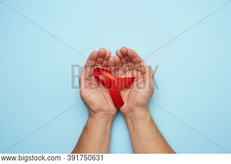 Male Hands Holds A Red Ribbon - A Symbol Of The Fight Against Disease Aids And Vasculitis, Blue Back